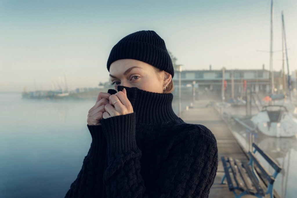 Eine Frau mit schwarzen Strickpullover und einer schwarzen Strickmütze hält sich den Pullover als Kälteschutz vor die Nase. Im Hintergrund ein Bootssteg.