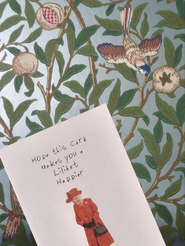 Vor einer kunstvollen Tapete mit einem Strauch und Vögeln wird eine Karte mit Queen Elisabeth hochgehalten. Die Beschriftung lautet: Hope this card makes you a lilibet happier.